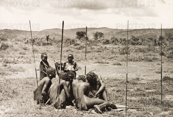 Young Maasai 'morani'. A group of young Maasai 'morani' (warriors) sit closely together on the ground, surrounded by their upright spears. An original caption comments that the group were employed as herdsman on a European settler's ranch. Kenya, circa 1935. Kenya, Eastern Africa, Africa.