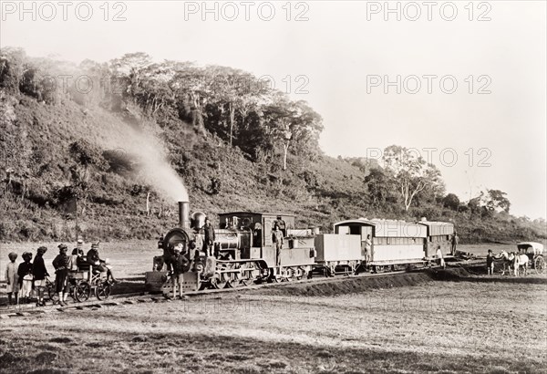 A steam train at Limuru. African railway staff gather around a stationary steam train on a track at Limuru. A European couple sit on a hand-operated rail cart in front of the train, with a team of African attendants behind them. Limuru, Kenya, 1903. Limuru, Central (Kenya), Kenya, Eastern Africa, Africa.