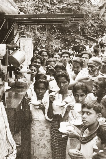 Drinking tea from a mobile kiosk. A group of people crowd around the hatch of a mobile kiosk, where an attendant offers drinks in cups and saucers. Ceylon (Sri Lanka), 1936. Sri Lanka, Southern Asia, Asia.