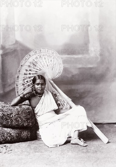 Studio portrait of a Ceylonian woman. Seated studio portrait of a Ceylonian woman in traditional dress. She leans against a cushion, propped up on one elbow and holding a giant, patterned fan in her other hand. She wears jewellery including bangles, necklaces, a bindi and a nose ring, and is dressed in a long sari over a 'choli' (blouse). Ceylon (Sri Lanka), circa 1900. Sri Lanka, Southern Asia, Asia.