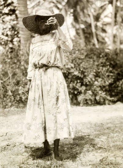 Grenadian woman smoking a pipe. Outdoors portrait of a Grenadian woman wearing a Western-style dress and a wide-brimmed sun hat, standing barefoot in the grass and smoking a pipe. Grenada, circa 1931. Grenada, Caribbean, North America .