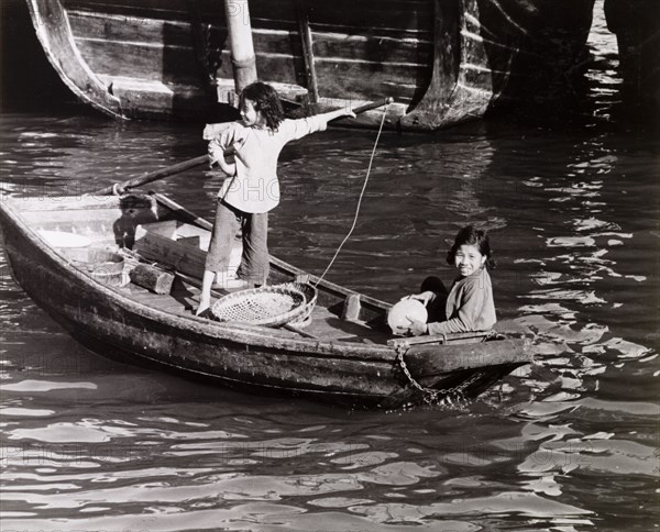 Chinese girls steer a sampan. Two girls travel aboard a scull-propelled sampan in Aberdeen Harbour, a vessel described in an original caption as their "family car". Aberdeen, Hong Kong, China, 1963. Aberdeen, Hong Kong, China, People's Republic of, Eastern Asia, Asia.