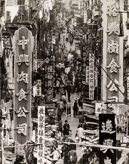 A busy side street in Hong Kong. Street traders vie for business on a city side street beneath a profusion of signs and advertisements written in Chinese script. Hong Kong, China, 1963. Hong Kong, Hong Kong, China, People's Republic of, Eastern Asia, Asia.