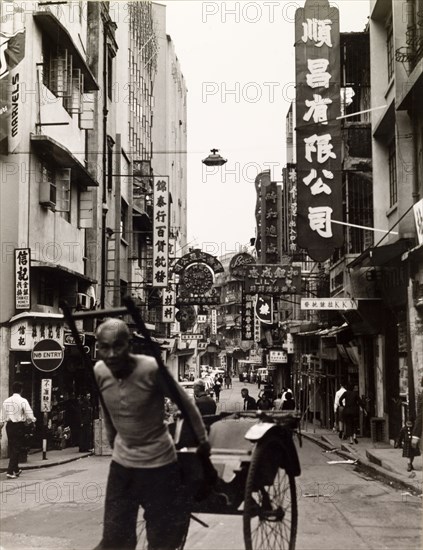Rickshaw driver in Hong Kong. A rickshaw driver pulls his empty carriage along a commerical street in Hong Kong. Signs and advertisements written in Chinese script stretch away into the distance. Hong Kong, China, 1963., Hong Kong, China, People's Republic of, Eastern Asia, Asia.