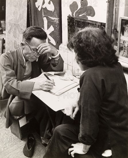 Letter-writing service on a Hong Kong street. A Chinese scribe sits at his street stall, concentrating as he writes a letter for a female customer. An original caption comments that he was "able write all kinds of Chinese and English letters, documents, applications, government and business forms". Hong Kong, China, 1963. Hong Kong, Hong Kong, China, People's Republic of, Eastern Asia, Asia.