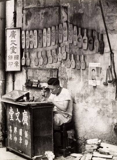 A mould maker, Hong Kong. A Chinese mould maker sits at his street stall, carving seals and cake moulds out of wood. Rows of completed moulds are strung up on the wall behind him. Hong Kong, China, 1963. Hong Kong, Hong Kong, China, People's Republic of, Eastern Asia, Asia.