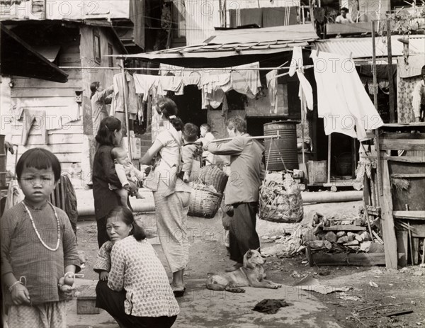 A squatter's settlement in Kowloon. Domestic scene in a Kowloon squatter's settlement. A woman hangs clothes on a makeshift washing line whilst others look after young children. A man walks right to left, carrying firewood hung from a pole across his shoulders. Kowloon, Hong Kong, China, 1963. Kowloon, Hong Kong, China, People's Republic of, Eastern Asia, Asia.