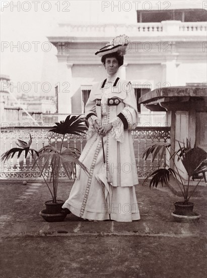 Minnie Murray in Edwardian dress. Full-length portrait of Minnie Murray, taken on a rooftop balcony in Calcutta. She stands between two pot plants, holding a parasol with both hands and wearing typical Edwardian attire including a formal dress with lace sleeves and a matching hat. 
Calcutta. Calcutta (Kolkata), India, 1906. Kolkata, West Bengal, India, Southern Asia, Asia.