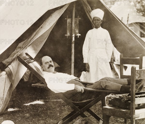 Taking a break at a camp site, India. James Murray, attended to by an Indian servant, reclines in a chair outside the entrance of a tent during a rest stop on a car trip from Calcutta (Kolkata) to Ranchi. India, circa 1916. India, Southern Asia, Asia.