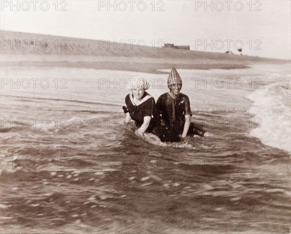 Minnie Murray and friend on a Puri beach. Minnie Murray (right) and a friend sit on a sandy beach in their swimming costumes, surrounded by the waves as they lap the shore. Puri, India, 1917. Puri, Orissa, India, Southern Asia, Asia.
