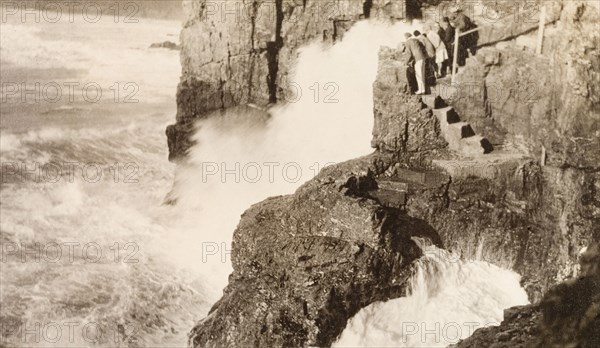 Watching the waves, Cornwall. A group of tourists stand on steps cut into a rocky outcrop on the north coast of Cornwall, watching the waves from the Atlantic Ocean crash into the rocks below. Perranporth, Cornwall, England, 1927., Cornwall, England (United Kingdom), Western Europe, Europe .