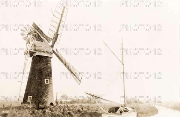 Sailing on the Norfolk Broads. A boat sails past a windmill as it travels along a river on the Norfolk Broads. Norfolk, England, 1930., Norfolk, England (United Kingdom), Western Europe, Europe .