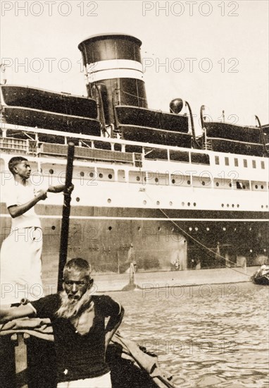 Ferry across the Hooghly River. A 'sarang' (senior crew member) ferries people across the Hooghly River on a dinghy to board a large steamship moored at Calcutta docks. Calcutta (Kolkata), India, circa 1931. Kolkata, West Bengal, India, Southern Asia, Asia.