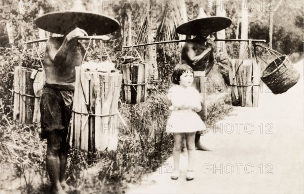 Transporting firewood, Kuala Lumpur. A young British girl visiting Kuala Lumpur stands between two Malaysian men carrying firewood hung from poles across their shoulders. Kuala Lumpur, Malay (Malaysia), 1931. Kuala Lumpur, Kuala Lumpur, Malaysia, South East Asia, Asia.