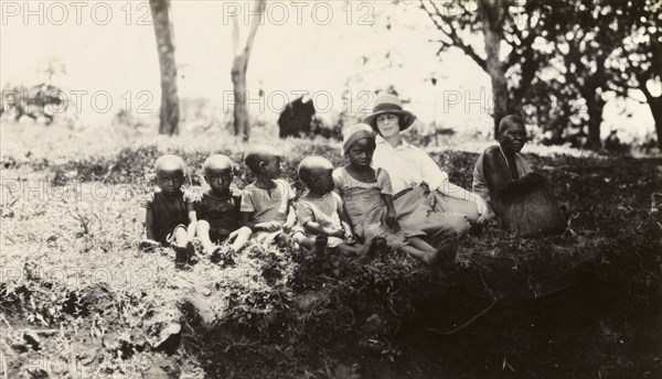 European woman with Chaga children. A European woman sits on a grassy bank with a Chaga woman and five young Chaga children. Tanganyika Territory (Tanzania), circa 1930. Tanzania, Eastern Africa, Africa.