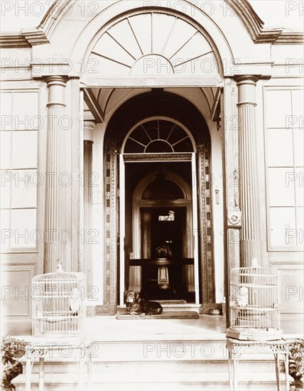 Porch of a grand, colonial house. The grand, pillared entrance to a colonial household. A pet dog rests on a mat in the porch, while two caged African grey parrots are positioned on either side of the doorway. St Helena, circa 1926. St Helena, Atlantic Ocean, Africa.