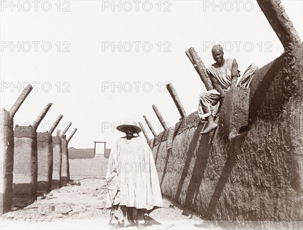 Emir's agent visits the site of a new school. A Nigerian man identified as 'Daudu, one of the Emir of Kano's agents' stands between mud walls at the construction site of a new school. A builder sits on the wall above him, smiling for the camera. Kano, Nigeria, 1914. Kano, Kano, Nigeria, Western Africa, Africa.