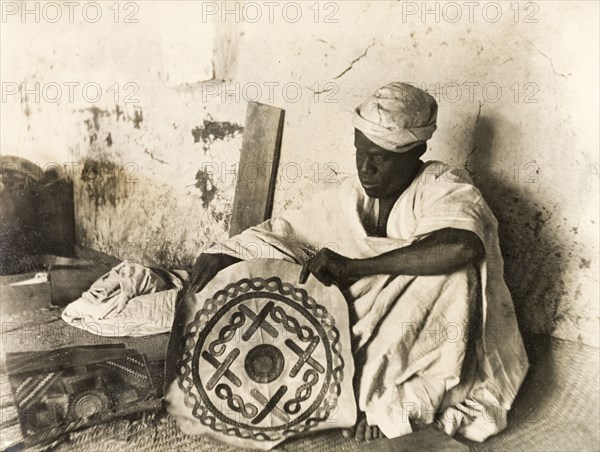 A Nigerian leatherworker. A Nigerian leatherworker sits in his workshop, holding up a decorative piece of leather that he has crafted. Probably Kano, Nigeria, circa 1915. Nigeria, Western Africa, Africa.