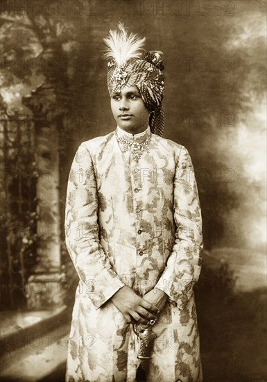 Portrait of a Maharajah. Studio portrait of an unidentified Maharajah dressed in ceremonial attire. He stands holding the hilt of a sword, wearing an embroidered 'sherwani' (overgarment) and a turban adorned with a feather and jewelled 'sarpech' (broach). He wears a ring on one hand and an intricate jewelled necklace around his collar. India, circa 1920., Maharashtra, India, Southern Asia, Asia.