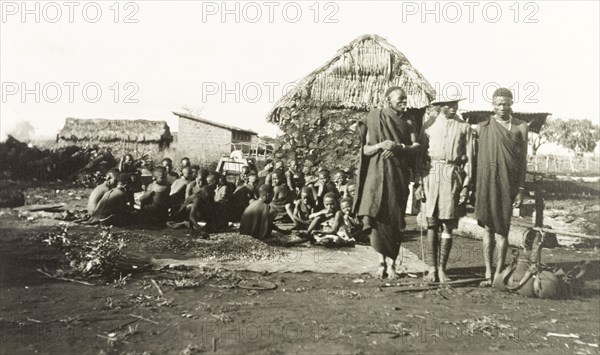 The Mission Boy'. A young African missionary, dressed in Western-style clothing and a solatopi hat, poses for the camera between two Kikuyu elders. A large group of Kikuyu women and children sit on the ground in the rural settlement behind them. Kenya, circa 1935. Kenya, Eastern Africa, Africa.