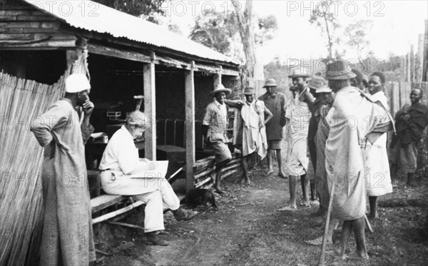 Pay day on a Kenyan farm. Kenyan farm hands gather around a European farmer's wife as she calculates their wages. Kenya, circa 1935. Kenya, Eastern Africa, Africa.