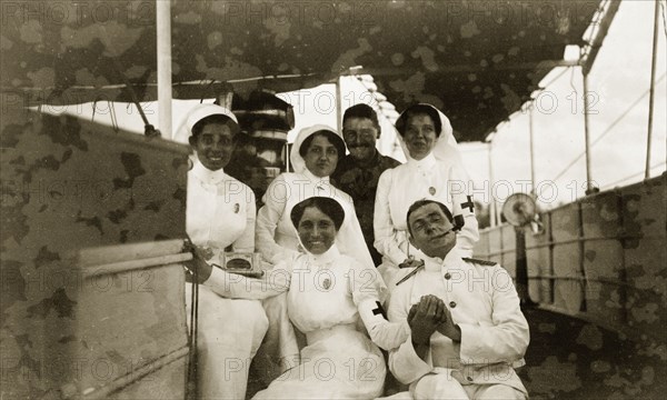 British officers with Indian nurses. Two British military officers pose on the deck of a ship with four Indian nurses. One of the men appears to be wearing a white naval uniform, whilst the other seems to be dressed in khaki army attire. Related images suggest the group were involved with the British offensive against German East Africa (Tanzania) during the First World War (1914-18). Probably Indian Ocean, circa 1914., Indian Ocean, Africa.