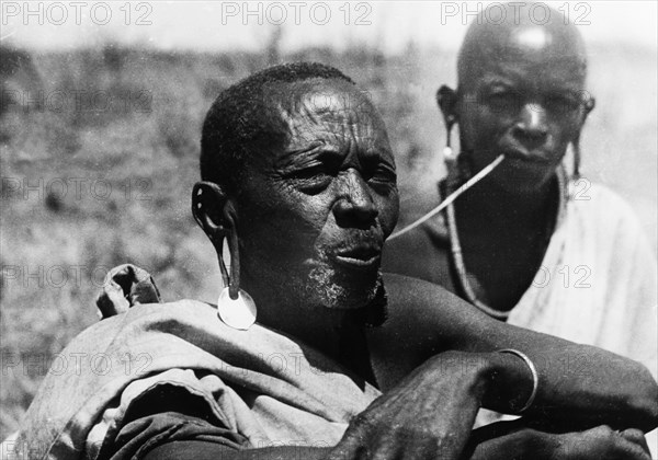 An elderly Kikuyu man. Portrait of an elderly Kikuyu man with a metal earring. Behind him sits a younger man, chewing on a long stalk of grass. Kenya, circa 1935. Kenya, Eastern Africa, Africa.
