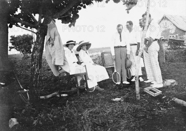 Tennis party at Kitimura Estate. Six European friends gather for a game of tennis at Kitimura Estate, the home of settler farmers Jos (standing with racquet) and Nellie Grant (left, seated). The Grants arrived in Kenya in 1912 to set up a coffee farm and were joined in 1914 by their seven year old daughter, Elspeth (later Huxley). Thika, Kenya, 1914. Thika, Nairobi Area, Kenya, Eastern Africa, Africa.