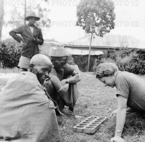 Elspeth Huxley plays a mancala game. Elspeth Huxley contemplates her next move as she plays a mancala game outdoors with two Kikuyu men. Njoro, Kenya, circa 1938. Njoro, Rift Valley, Kenya, Eastern Africa, Africa.