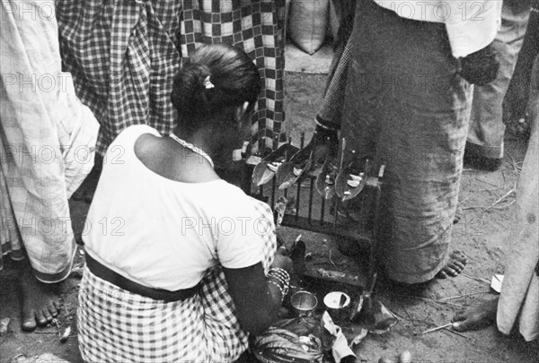 Short-eats' in Ceylon. A female market trader squats at her stall, apparently preparing some sort of 'short-eats' (snacks) wrapped in leaves. Ceylon (Sri Lanka), circa 1935. Sri Lanka, Southern Asia, Asia.
