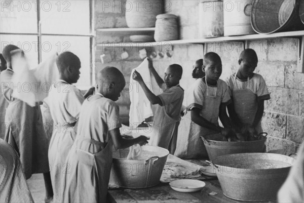 Laundry at a mission school. Kikuyu boys dressed in Western-style uniforms and aprons wash clothes in metal tubs inside a stone-walled laundry room. They are probably students of a Christian mission school, possibly the Church of Scotland mission school in south Nyeri. Kenya, circa 1936. Kenya, Eastern Africa, Africa.