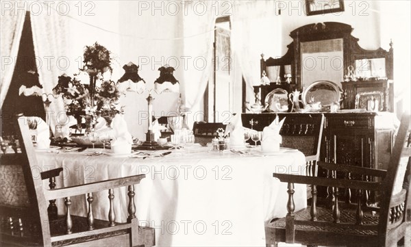 Governor's dining room at Edakkara lodge. Interior shot of a dining room in the bungalow at Edakkara hunting lodge, taken during the stay of Sir Arthur Lawley, the Governor of Madras. In the centre of the room stands a table set with fine china and flowers, with a cabinet displaying silverware standing against the far wall. Edakkara, Malabar District (Kerala), India, March 1908., Kerala, India, Southern Asia, Asia.