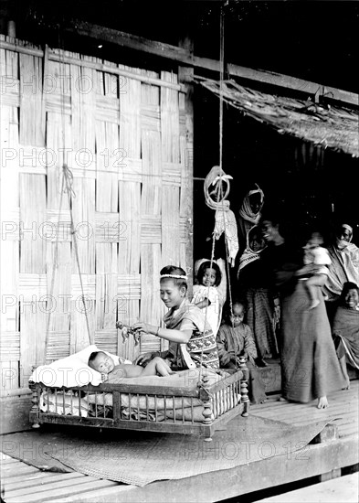 Baby in a wooden cradle, Burma. A Burmese lady amuses a baby with a toy as it lies in a hanging cradle on the decked veranda of a domestic hut. Burma (Myanmar), circa 1890. Burma (Myanmar), South East Asia, Asia.