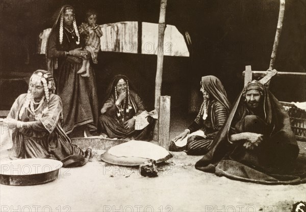 Palestinian bedouins at their tent. Four bedouin women, all dressed in long robes with headscarfs and beaded headpieces, one holding an infant, chat as they prepare food at the entrance to their tent in the company of an elderly man. British Mandate of Palestine (Middle East), circa 1942., Middle East, Asia.