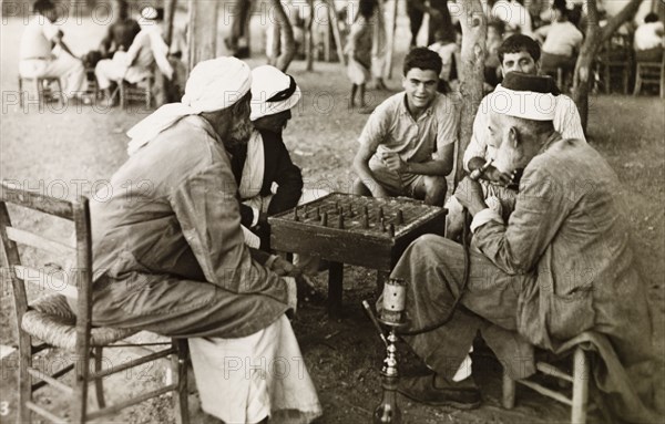 Socializing outside a cafe, Palestine. A group of Palestinian men sit around a table outside a cafe, playing a board game and smoking a water pipe. British Mandate of Palestine (Middle East), circa 1942., Middle East, Asia.