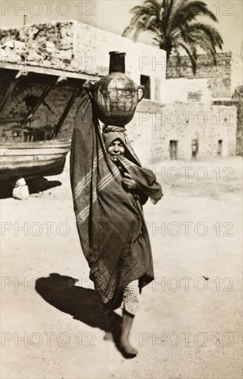 Palestinian woman collecting water. A Palestinian woman in traditional Arabic dress balances a clay pot full of water on her head as she walks along a village street. British Mandate of Palestine (Middle East), circa 1942., Middle East, Asia.