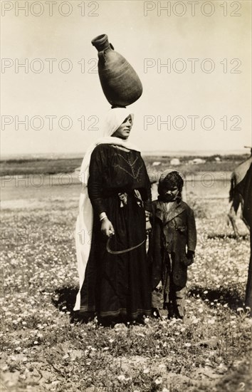 Bedouin woman carrying water pot. A bedouin woman stands with a young child in the Palestinian countryside, balancing a large clay pot on her head at a jaunty angle. British Mandate of Palestine (Middle East), circa 1942., Middle East, Asia.