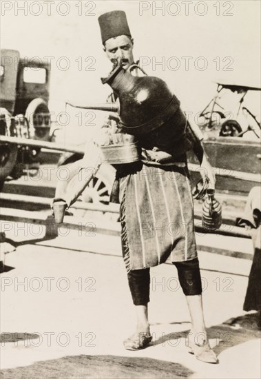 Cold drink seller, Palestine. A street peddler with a large jug strapped to his chest stands by the roadside plying for trade, leaning over to one side to pour a drink from the jug into a cup. British Mandate of Palestine (Middle East), circa 1942., Middle East, Asia.