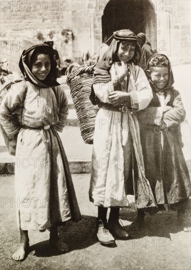 Child basket carriers, Palestine. Three Palestinian children pose on a city street, each carrying a large basket on their back with the aid of a strap attached to their head. British Mandate of Palestine (Middle East), circa 1942., Middle East, Asia.