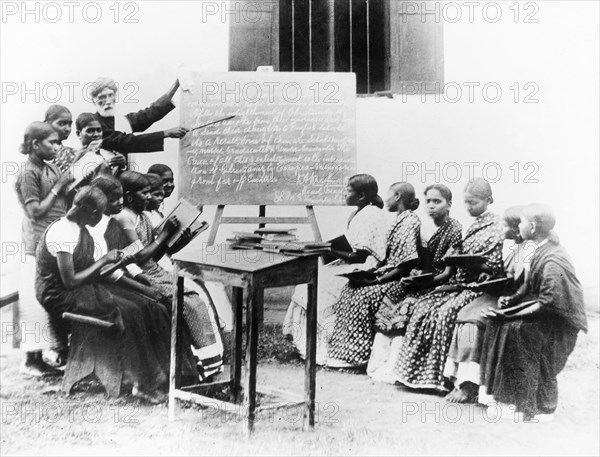 English lesson at a mission school, India. An Indian teacher points to a blackboard as he gives an English lesson outdoors at a school run by the London Missionary Society. He is surrounded by a class of Indian students who conscientiously copy text from the blackboard onto their slates. India, circa 1930. India, Southern Asia, Asia.