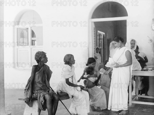 Queuing outside a mission hospital, India. Indian women and girls sit on a bench as they queue for medical attention outside a hospital run by the London Missionary Society. India, circa 1930. India, Southern Asia, Asia.