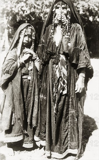 Portrait of two female 'bedouins'. Full-length portrait of an Arab woman and girl, identified in an original caption as ?bedouin(s)?. They wear long embroidered 'thobs', typical of Palestinian dress, with headscarves trailing down their backs and elaborate coin jewellery around their faces. British Mandate of Palestine (Middle East), circa 1938., Middle East, Asia.
