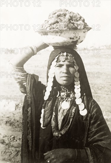 Portrait of a bedouin woman. Half-length portrait of a bedouin woman carrying a large bowl on her head. She wears a traditional Palestinian 'thob' and a headscarf fringed with coins. British Mandate of Palestine (Middle East), circa 1938., Middle East, Asia.