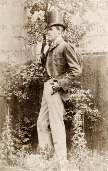 James Murray on his 21st birthday. Full-length portrait of James Murray, taken on his 21st birthday. He smokes a cigarette as he leans against a garden fence, dressed in smart Victorian attire including a tall top hat. England, 11 June 1891. England (United Kingdom), Western Europe, Europe .