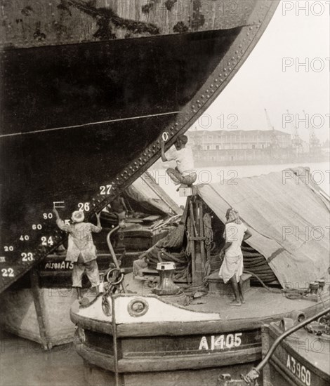 How to paint a ship when you don't have a ladder'. Three dock workers on a river barge attempt to paint measurements onto the hull of a large steamship without the use of a ladder. Calcutta (Kolkata), India, 1931. Kolkata, West Bengal, India, Southern Asia, Asia.