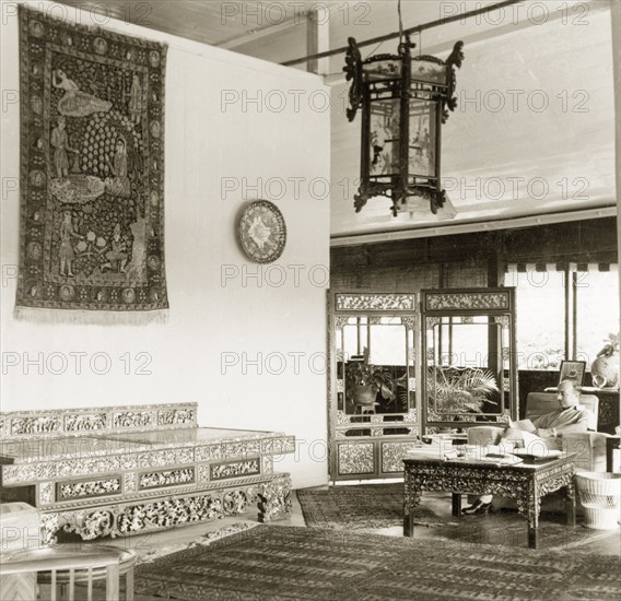 Asian artefacts in a colonial drawing room, British Malaya. Dr Reid Tweedie sits reading in the drawing room of his stilted bungalow. The room is furnished with Asian-style furniture, tapestries and ornaments. Sungei Siput near Ipoh, British Malaya (Malaysia), August 1940. Ipoh, Perak, Malaysia, South East Asia, Asia.