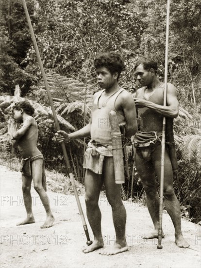Orang Asli with blow pipes. Two Orang Asli men stand on a rural road, each holding a long blowpipe made from a hollow bamboo tube. These pipes, still used today, are loaded with poisoned darts with which to kill monkeys and birds in the rainforests of Malaysia. British Malaya (Malaysia), 1940. Malaysia, South East Asia, Asia.