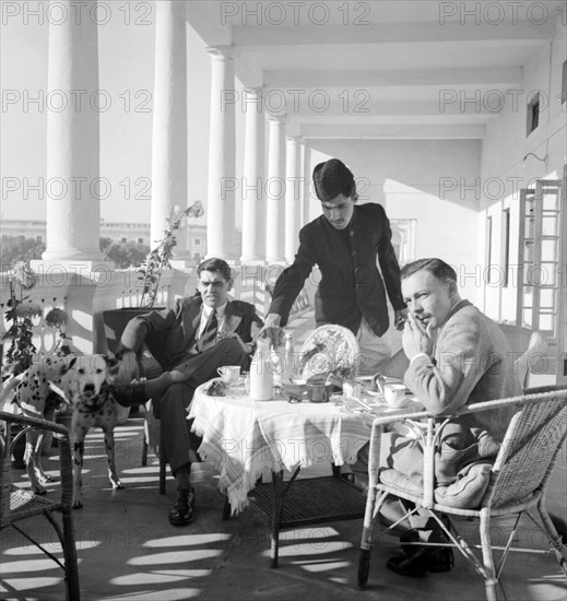 Tea on the veranda, India. Two European men take tea and breakfast on the pillared veranda of a colonial building, attended to by an Indian servant and accompanied by a pet Dalmation dog. India, circa 1935. India, Southern Asia, Asia.
