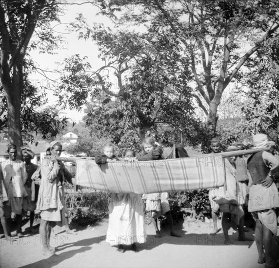 Children travelling in a sedan chair. An Indian ayah (nursemaid) watches over two European children as they travel in a sedan chair carried by Indian servants. India, circa 1935. India, Southern Asia, Asia.