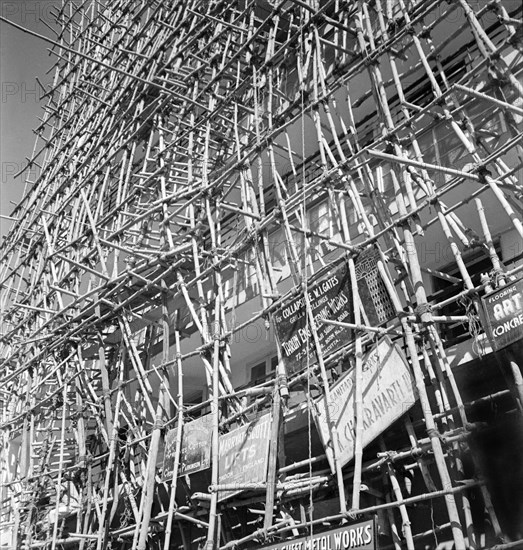 Bamboo scaffolding with adverts, Calcutta. A tall building is encased in bamboo scaffolding as it undergoes construction. A number of signs hang from the canes, advertising construction companies and engineering works. Calcutta (Kolkata), India, circa 1940. Kolkata, West Bengal, India, Southern Asia, Asia.